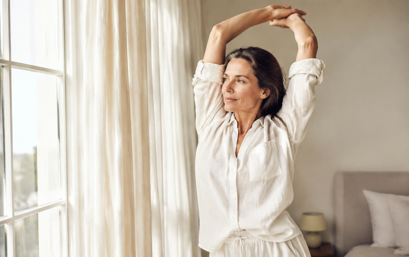 Woman in her late 40s stretching by a bright window